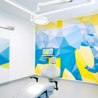 Behandlungsraum bei MKG Plus mit kunstvoll gestalteter Wand im Hintergrund. Sie zeigt einen großen, blauen Elefanten vor gelbem Hintergrund.