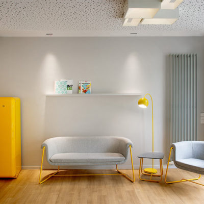 Blick in einen Wartebereich der Praxis MKG Plus in Münster. Eine moderne Couch, ein Sessel und ein gelber Kühlschrank sind zu sehen. Der Boden hat einen Holzbelag.
