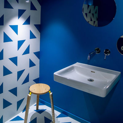 Zu sehen ist ein modernes Waschbecken. An einer dunkelblauen Wand hängt ein runder Spiegel. Links neben dem Waschbecken steht ein Holzhocker. Die linke Wand und der Boden sind kunstvoll in weiß und blau gefliest.