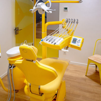 Behandlungsraum mit weißen Wänden und Holzfußboden. Der Behandlungsstuhl, die Geräte und ein zus. Stuhl sind gelb.