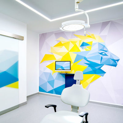 Behandlungsraum bei MKG Plus mit kunstvoll gestalteter Wand im Hintergrund. Sie zeigt einen großen, blauen Bären vor gelbem Hintergrund.