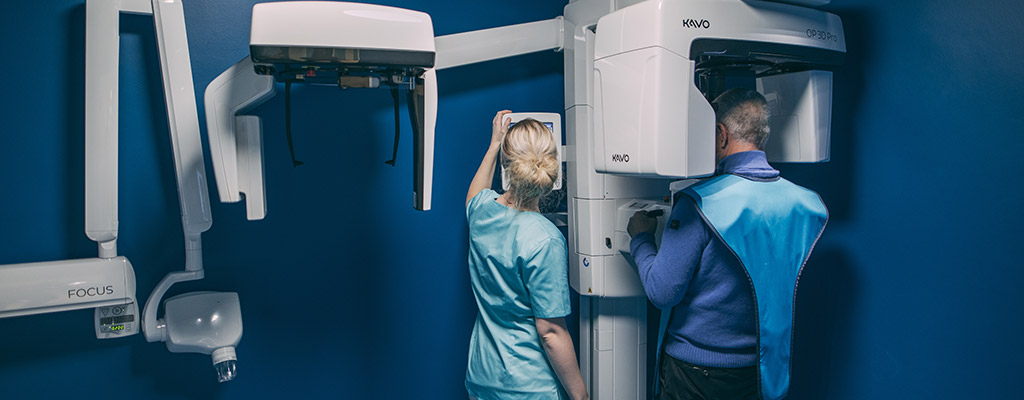 Ein grauhaariger Mann steht mit dem Rücken zur Kamera in einem 3D-Röntgengerät. Eine blonde Frau, ebenfalls mit dem Rücken zur Kamera, bedient ein Display. Das mannshohe Röntgengerät steht vor einer dunkelblauen Wand.
