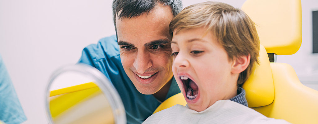 Ein Junge sitzt in einem gelben Behandlungsstuhl und schaut sich im Handspiegel seine Zähne an.