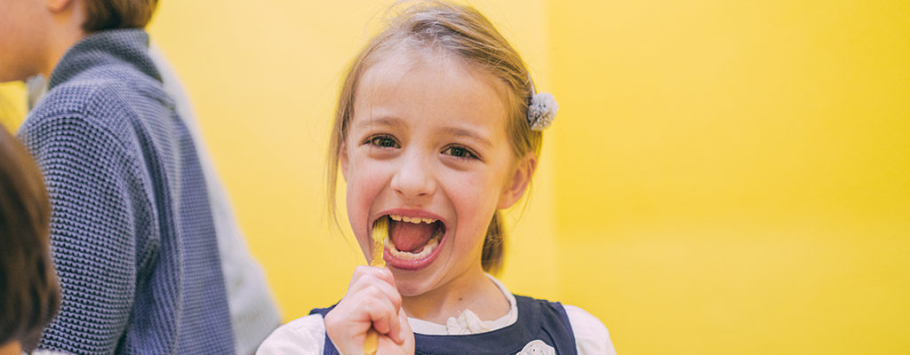 Ein blondes Mädchen steht von einer gelben Wand, blickt in die Kamera und putzt mit einer gelben Zahnbürste ihre Zähne.