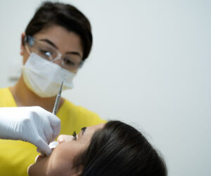 Das Bild zeigt Achin Farec, eine zahnmedizinische Prophylaxeassistentin und zahnmedizinische Fachangestellte, bei der Durchführung einer professionellen Zahnreinigung (PZR) in Münster. Sie trägt eine Schutzbrille, eine Maske und gelbe medizinische Kleidung. Achin Farec hält ein zahnärztliches Instrument und arbeitet an den Zähnen einer Patientin, die auf dem Zahnarztstuhl liegt.