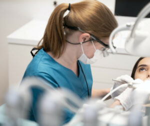 Das Bild zeigt eine zahnmedizinische Fachkraft bei der Arbeit an einer Patientin. Die Fachkraft trägt blaue medizinische Kleidung, eine Lupenbrille und eine Maske. Sie hält zahnärztliche Instrumente in den Händen und beugt sich über die Patientin, die auf einem Zahnarztstuhl liegt. Im Hintergrund sind zahnärztliche Geräte und eine weiße Schublade zu sehen.