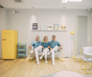 Das Bild zeigt Dr. Ponelis, Dr. Ellermann und Dr. Kedir, die auf einer Bank im Wartebereich ihrer Zahnarztpraxis sitzen. Alle drei tragen blaue medizinische Kleidung. Der Wartebereich ist modern gestaltet, mit einem gelben Kühlschrank, einem blauen Rollwagen und einer gelben Stehlampe. Im Hintergrund ist ein Regal mit Büchern und Dekorationen an der Wand angebracht. Der Boden ist aus hellem Holz.