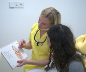 Das Bild zeigt eine Zahnmedizinische Prophylaxeassistentin in gelber medizinischer Kleidung, die einer Patientin eine Illustration aus einem Lehrbuch erklärt. Die Prophylaxeassistentin hält das Buch in der linken Hand und zeigt auf ein Diagramm, das die Struktur eines Zahns und des Zahnfleisches darstellt. Die Patientin sitzt neben ihr und hört aufmerksam zu. Die Prophylaxeassistentin trägt eine zahnärztliche Lupenbrille um den Hals und ihre Gesichtsmaske ist unter das Kinn gezogen, um besser erklären zu können.