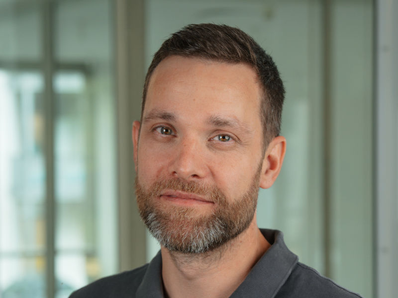 Das Bild zeigt David Roters, Zahntechnikermeister bei Zahnärzte MKG+ Münster. Er trägt ein graues Poloshirt und lächelt freundlich in die Kamera. David Roters hat kurze, braune Haare und einen gepflegten Vollbart.