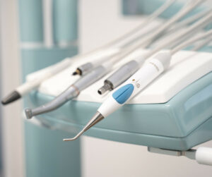 Das Bild zeigt eine Nahaufnahme einer Ablage für zahnärztliche Instrumente. Auf der Ablage befinden sich mehrere zahnmedizinische Werkzeuge, darunter ein Ultraschall-Scaler mit einer blauen und weißen Oberfläche sowie weitere Handstücke für verschiedene zahnärztliche Behandlungen. Die Instrumente sind ordentlich angeordnet und die Ablage ist in einem hellblauen und weißen Farbschema gestaltet, das einen sauberen und modernen Eindruck vermittelt. Im Hintergrund ist ein Teil der Zahnarztpraxis zu sehen, ebenfalls in hellen und freundlichen Farben gehalten.