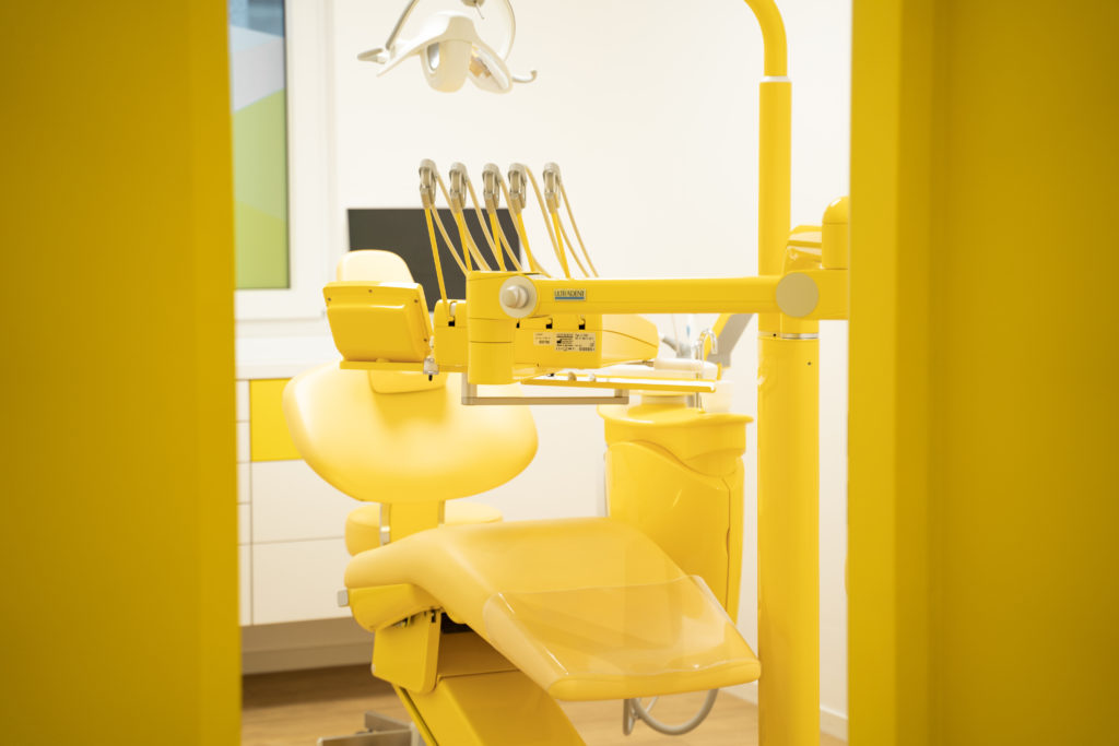 Behandlungsstuhl beim Zahnarzt - Die Wände des Behandlungsraums sind weiß, der Behandlungsstuhl und die Behandlungsgeräte sind knallgelb.