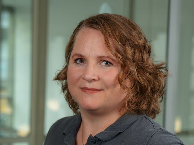 Das Bild zeigt Alexandra Stewart, Zahntechnikermeisterin bei Zahnärzte MKG+ Münster. Sie trägt ein graues Poloshirt und lächelt freundlich in die Kamera. Alexandra Stewart hat schulterlange, hellbraune, wellige Haare.