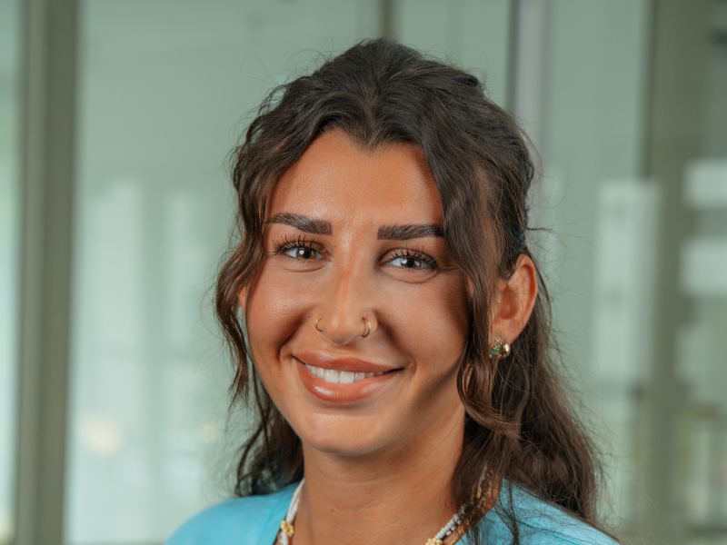 Das Bild zeigt Emilija Boskovic, eine zahnmedizinische Fachangestellte bei den Zahnärzten MKG+ Münster. Sie trägt eine blaue Arbeitskleidung und lächelt freundlich in die Kamera. Emilija hat schulterlanges, braunes Haar mit sanften Wellen.
