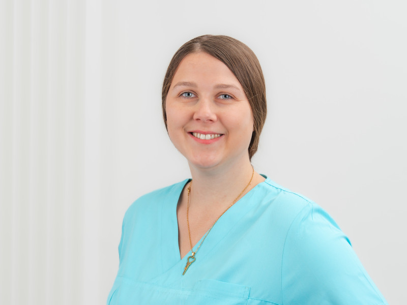 Das Bild zeigt Lena Dütting, eine Zahnmedizinische Fachangestellte.Sie trägt hellblaue medizinische Kleidung und lächelt freundlich in die Kamera. Lena Dütting trägt ihre glatte, hellbraune Haare zu einem Zopf gebunden.
