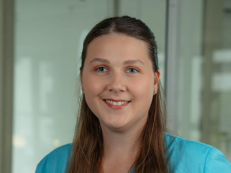 Das Bild zeigt Lena Dütting, eine Zahnmedizinische Fachangestellte.Sie trägt hellblaue medizinische Kleidung und lächelt freundlich in die Kamera. Lena Dütting hat lange, glatte, hellbraune Haare.
