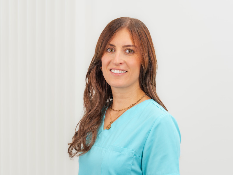 Michelle Speth, Zahnmedizinische Fachangestellte und Teamleitung Assistenz, steht leicht seitlich vor einem hellen, strukturierten Hintergrund und blickt freundlich in die Kamera. Sie trägt türkisfarbene medizinische Berufskleidung. Ihr langes, welliges Haar ist offen gestylt und fällt über die Schultern. Dezente Accessoires wie goldfarbene Halsketten und kleine runde Ohrringe ergänzen ihr gepflegtes Erscheinungsbild.