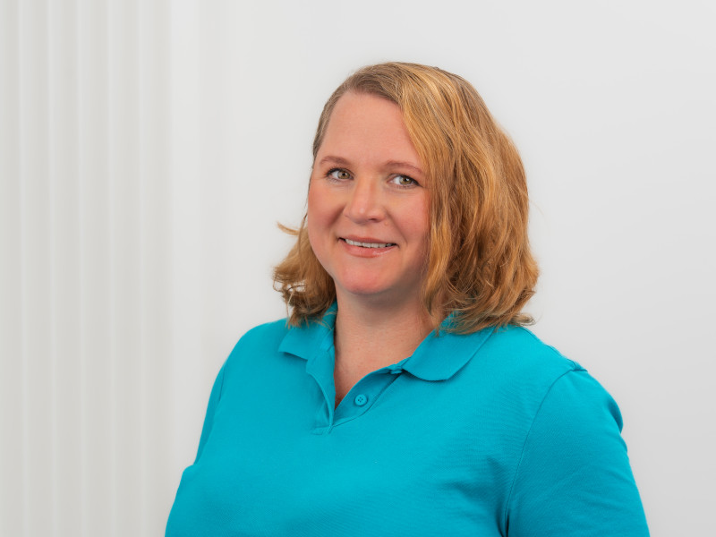 Das Bild zeigt Alexandra Stewart, Zahntechnikermeisterin bei Zahnärzte MKG+ Münster. Sie trägt ein blaues Poloshirt und lächelt freundlich in die Kamera. Alexandra Stewart hat schulterlange, hellbraune, wellige Haare.