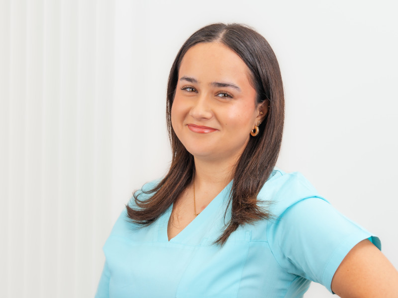 Defne Kurt, Auszubildende zur Zahnmedizinischen Fachangestellten, steht vor einem hellen Hintergrund mit dezentem Streifenverlauf und blickt mit einem selbstbewussten Lächeln in die Kamera. Sie trägt türkisfarbene medizinische Arbeitskleidung, ihr glattes, dunkles Haar ist offen und fällt bis auf die Schultern. Runde, goldene Ohrringe und eine feine Halskette ergänzen ihr gepflegtes Erscheinungsbild. Die Ausstrahlung wirkt freundlich und zugewandt.