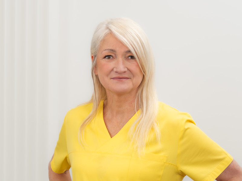 Das Bild zeigt Margarete Lolaj, eine Zahnmedizinische Fachangestellte im Bereich Prophylaxe. Sie trägt gelbe medizinische Kleidung und lächelt freundlich in die Kamera. Margarete Lolaj hat lange, glatte, blonde Haare und braune Augen.