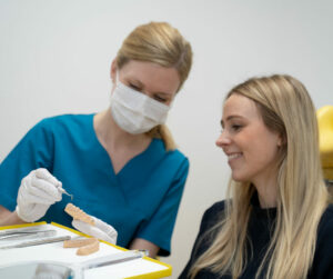 Das Bild zeigt Dr. Sophia Ellermann in blauer medizinischer Kleidung und einer Gesichtsmaske, wie sie einer Patientin etwas erklärt. Dr. Ellermann hält ein Zahnmodell in der Hand und verwendet ein zahnärztliches Instrument, um etwas darauf zu zeigen. Die Patientin, eine junge Frau mit langen blonden Haaren, sitzt lächelnd neben ihr und hört aufmerksam zu. Auf der Ablage vor ihnen liegen weitere zahnärztliche Instrumente. Die Szene findet in einer modernen und hellen Zahnarztpraxis statt, die eine freundliche und professionelle Atmosphäre ausstrahlt.