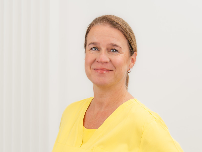 Tanja Zengeley, Zahnmedizinische Fachangestellte mit den Aufgabenbereichen Prophylaxe und Verwaltung, steht vor einem weißen Hintergrund mit dezenten senkrechten Strukturen. Sie trägt gelbe medizinische Arbeitskleidung, hat mittelblondes, zurückgebundenes Haar und silberne Ohrringe. Ihr Gesichtsausdruck ist freundlich und ruhig, die Haltung offen und professionell.