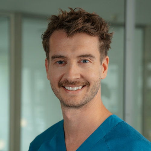 Das Bild zeigt Dr. Julius Balke, einen Zahnarzt in Münster. Er hat kurze, braune Haare und lächelt freundlich in die Kamera. Dr. Balke trägt eine blaue medizinische Kleidung. Der Hintergrund ist hell und schlicht gehalten.
