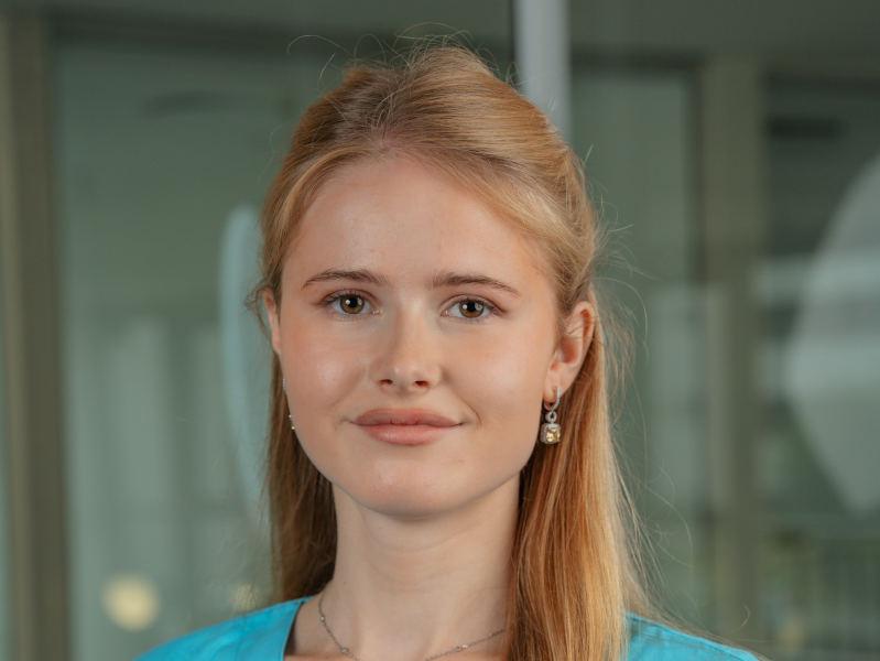 Das Bild zeigt Katharina Kraft, eine Auszubildende zur Zahnmedizinischen Fachangestellten (ZFA) bei MKG+ Zahnärzte Münster. Sie trägt hellblaue medizinische Kleidung und lächelt freundlich in die Kamera. Katharina Kraft hat lange, glatte, blonde Haare und braune Augen. Im Hintergrund sind weiße Wände zu sehen, was eine helle und einladende Atmosphäre schafft.