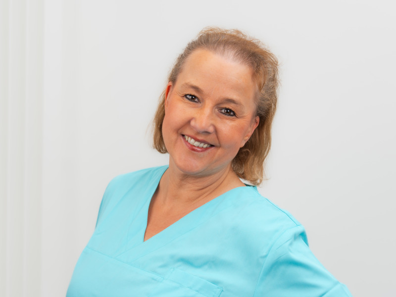 Lächelndes Porträt von Birgit Fohrmann, Technische Sterilisationsassistentin bei der Zahnarztpraxis MKG Plus in Münster. Sie trägt türkisfarbene Arbeitskleidung und schaut freundlich in die Kamera. Ihr rötlich-blondes Haar ist schulterlang und locker zurückgekämmt.