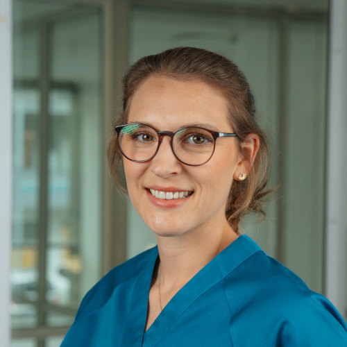 Porträtaufnahme der Zahnärztin Carla Borchert in den Räumen der Zahnarztpraxis MKG Plus in Münster. Sie steht vor einer Glaswand mit unscharfem Hintergrund, trägt eine moderne, blaue Arbeitskleidung und blickt freundlich in die Kamera. Carla Borchert hat hellbraune, schulterlange Haare, die locker nach hinten gebunden sind, trägt eine runde Brille mit dunklem Rahmen und dezente Ohrringe. Ihr Ausdruck ist ruhig und sympathisch – sie wirkt professionell und zugewandt.