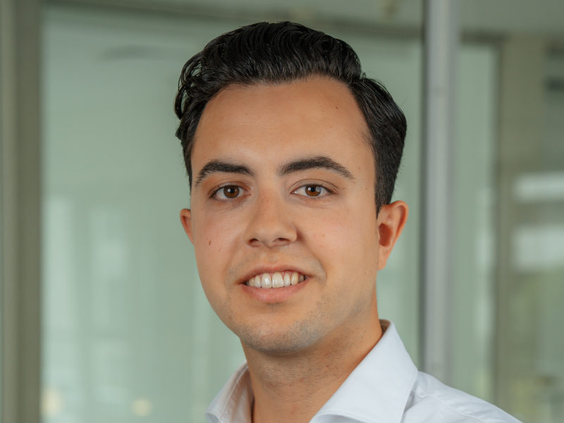 Porträtaufnahme von Filipe Henrique, Leiter des Personalbereichs in der Verwaltung der Zahnklinik MKG Plus in Münster. Er trägt ein weißes Hemd und blickt mit einem freundlichen, professionellen Ausdruck in die Kamera. Sein dunkles Haar ist sauber gestylt und zur Seite frisiert. Der Hintergrund besteht aus großen Glasflächen und wirkt modern und hell, ist jedoch unscharf, um den Fokus auf die Person zu legen.