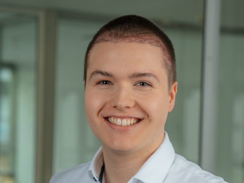 Freundliches Porträt von Jan Luca Grüneberg-Nolte, Mitarbeiter im Bereich Empfang und Verwaltung der Zahnklinik MKG Plus in Münster. Er trägt ein helles Hemd und lächelt offen in die Kamera. Sein kurzes, dunkelblondes Haar ist ordentlich frisiert. Der Hintergrund zeigt eine moderne, helle Glasarchitektur innerhalb der Praxis und ist leicht unscharf, wodurch der Fokus klar auf dem Gesicht liegt.
