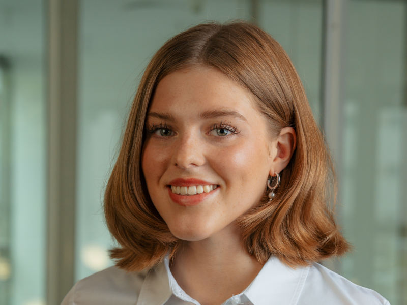 Porträt von Lilian Kammel, zahnmedizinische Fachangestellte im Bereich Empfang der Zahnklinik MKG Plus in Münster. Sie trägt eine weiße Bluse und hat kinnlanges, rötlich-blondes Haar, das leicht gewellt ist. Lilian lächelt herzlich in die Kamera. Sie trägt dezenten Ohrschmuck in Form kleiner silberner Creolen. Der Hintergrund zeigt eine moderne, lichtdurchflutete Glasarchitektur in weichem Fokus.