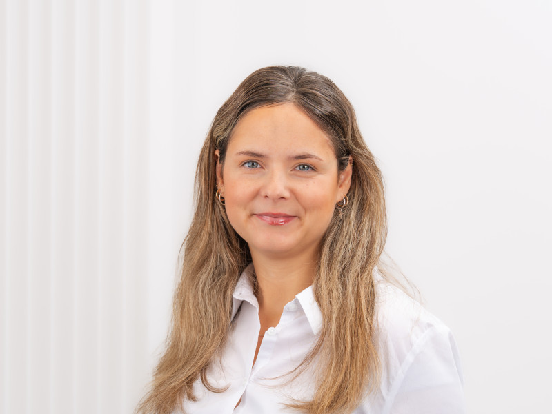 Porträt von Marlene dos Reis Goncalves, zahnmedizinische Fachangestellte in der Verwaltung der Zahnklinik MKG Plus in Münster. Sie trägt eine weiße Arbeitsbluse und hat langes, gewelltes, mittelblondes Haar. Marlene lächelt in die Kamera und strahlt dabei Offenheit und Freundlichkeit aus..