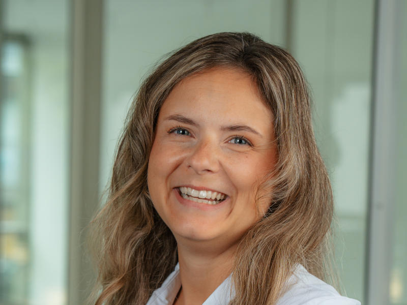 Fröhliches Porträt von Marlene dos Reis Goncalves, zahnmedizinische Fachangestellte in der Verwaltung der Zahnklinik MKG Plus in Münster. Sie trägt eine weiße Arbeitsbluse und hat langes, gewelltes, mittelblondes Haar. Marlene lächelt herzlich in die Kamera und strahlt dabei große Offenheit und Freundlichkeit aus. Der Hintergrund ist unscharf und zeigt eine helle Glasfront innerhalb der Praxisräume.