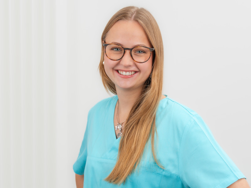 Franziska Hartung, Auszubildende zur Zahnmedizinischen Fachangestellten, steht vor einem hellen, weißen Hintergrund mit dezenten senkrechten Linien. Sie hat langes, glattes, blondes Haar und trägt eine Brille mit rundem Rahmen. Gekleidet ist sie in hellblauer medizinischer Arbeitskleidung. Sie lächelt freundlich in die Kamera, ihre Haltung wirkt selbstbewusst und offen.