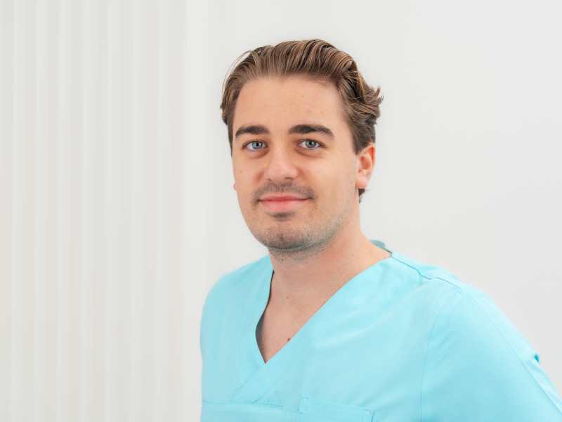 Henry Mehnert, Werkstudent in der Zahnarztpraxis, steht vor einem weißen Hintergrund mit senkrechten Linienstrukturen. Er trägt ein hellblaues Oberteil medizinischer Berufskleidung mit V-Ausschnitt. Sein braunes Haar ist zurückgekämmt, sein Blick ist ruhig und freundlich, und er lächelt dezent in die Kamera.