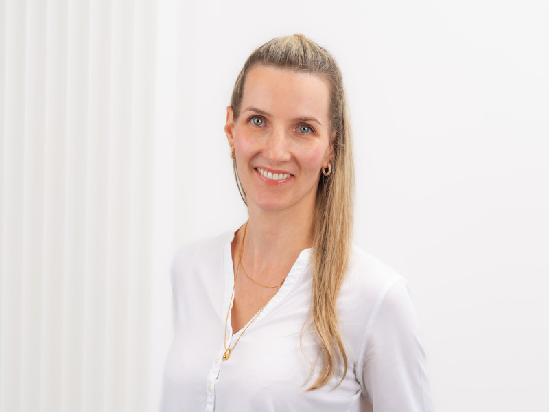 Linda Haase, Zahnmedizinische Fachangestellte in der Verwaltung, steht vor einem hellen, weißen Hintergrund mit dezenten senkrechten Strukturen. Sie hat langes, glattes, blondes Haar, das teilweise zurückgebunden ist, und trägt eine weiße Bluse sowie dezenten Schmuck in Gold. Ihr Gesichtsausdruck ist freundlich und offen, die Haltung aufrecht und zugewandt.