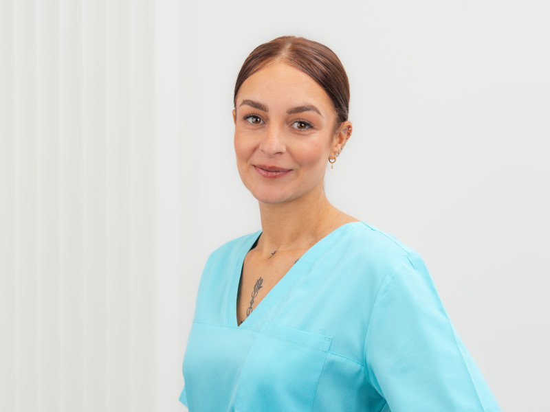 Melanie Stoever, Zahnmedizinische Fachangestellte, steht leicht seitlich zur Kamera und lächelt dezent. Sie trägt türkisfarbene medizinische Arbeitskleidung, hat dunkelbraunes, streng zurückgebundenes Haar und dezenten Goldschmuck. Der V-Ausschnitt ihrer Kleidung macht ein kleines Tattoo sichtbar. Der helle Hintergrund mit feiner Struktur unterstreicht ihre ruhige und freundliche Ausstrahlung.