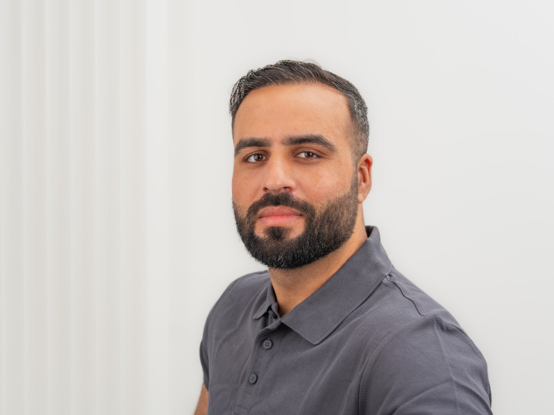Muqdad Murad, Auszubildender im Zahntechnikerhandwerk, blickt ruhig und selbstbewusst in die Kamera. Er trägt ein dunkelgraues Poloshirt, hat einen gepflegten Vollbart und kurz geschnittenes, dunkles Haar. Der Hintergrund ist schlicht weiß mit einem angedeuteten Strukturverlauf, wodurch die Person klar im Fokus steht. Die Atmosphäre wirkt professionell und seriös.