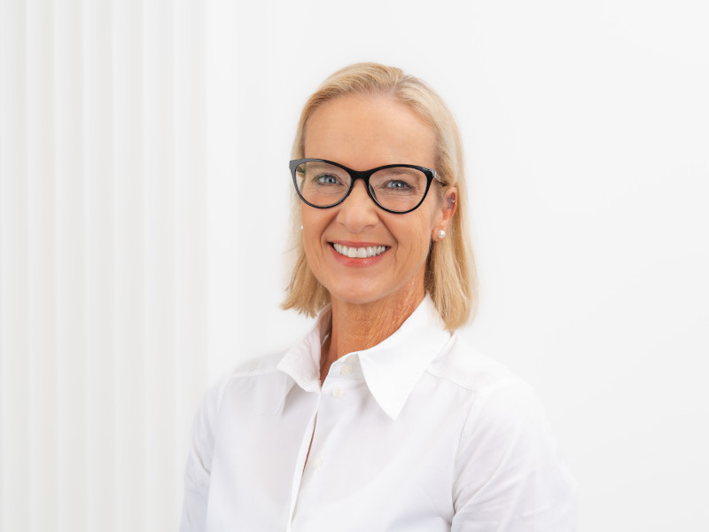 Ramona Wolf, Zahnmedizinische Fachangestellte mit Schwerpunkt Prophylaxe und Verwaltung, steht vor einem hellen, strukturierten Hintergrund. Sie trägt eine weiße Bluse, dazu Perlenohrringe und eine markante dunkle Brille. Ihr blondes Haar ist kinnlang geschnitten und ordentlich frisiert. Mit einem offenen Lächeln blickt sie direkt in die Kamera.