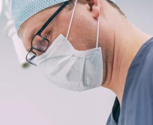 Weisheitszahnentfernung bei MKG Plus in Münster durch erfahrenen MKG-Chirurgen Nahaufnahme eines MKG-Chirurgen bei einer Weisheitszahnentfernung in der Zahnklinik MKG Plus in Münster. Der Facharzt trägt blaue OP-Kleidung, eine helle OP-Haube, schwarze Brille und einen Mundschutz. Sein Blick ist konzentriert nach unten auf das Behandlungsfeld gerichtet. Der helle, sterile Hintergrund unterstreicht die ruhige und präzise Atmosphäre des chirurgischen Eingriffs.