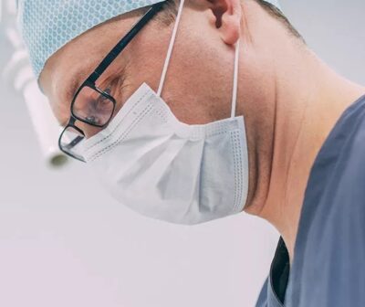 Nahaufnahme eines MKG-Chirurgen bei einer Weisheitszahnentfernung in der Zahnklinik MKG Plus in Münster. Der Facharzt trägt blaue OP-Kleidung, eine helle OP-Haube, schwarze Brille und einen Mundschutz. Sein Blick ist konzentriert nach unten auf das Behandlungsfeld gerichtet. Der helle, sterile Hintergrund unterstreicht die ruhige und präzise Atmosphäre des chirurgischen Eingriffs.